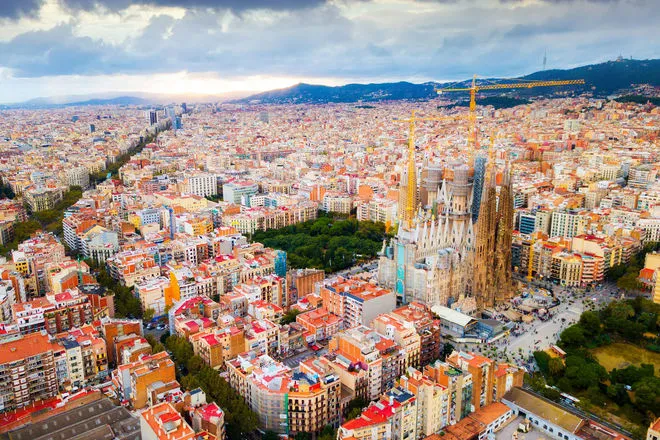 A panoramic view of Barcelona, Spain, featuring iconic landmarks such as the Sagrada Família and Park Güell against a backdrop of the cityscape and the Mediterranean Sea. The vibrant city is alive with culture, history, and architectural beauty.