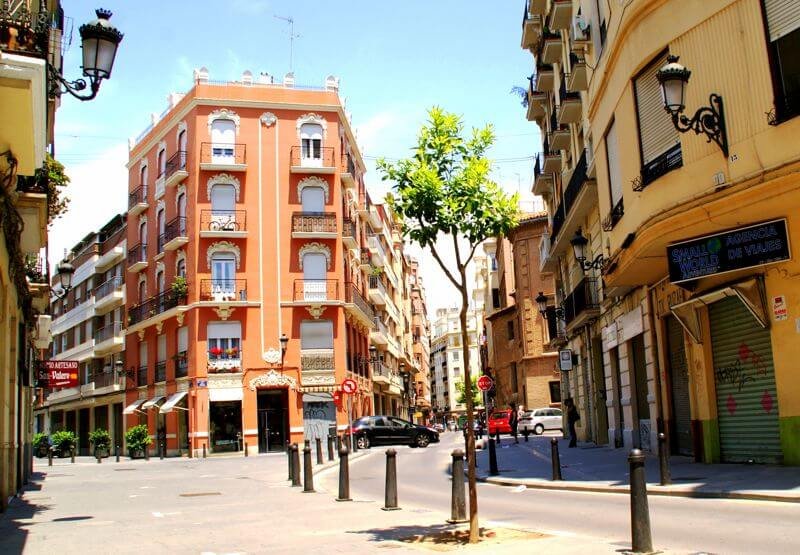 A vibrant street scene in the Ruzafa neighborhood of Valencia, Spain, showcasing colorful buildings, outdoor cafes, and locals enjoying the lively atmosphere. The area is known for its trendy vibe, artistic flair, and blend of traditional and modern culture.